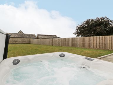A bubbling hot tub in a fenced backyard with grass and stone buildings in the background at The Byre in St Brides Major
