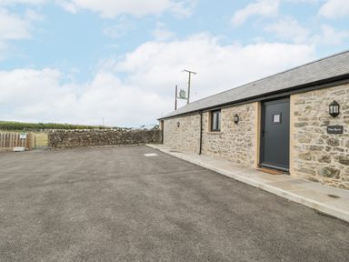 An exterior view of a stone building with a black door and small windows next to a paved area and a stone wall at The Byre in St Brides Major