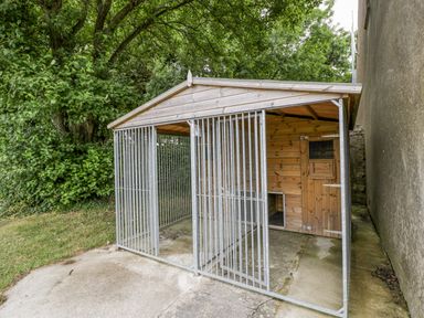 An outdoor kennel with metal bars and a wooden shelter next to a wall and trees at The Byre in St Brides Major