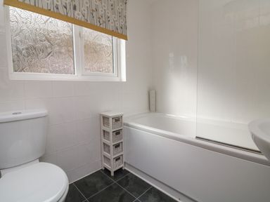 A bathroom with a toilet a bathtub a small white storage unit and a window with patterned glass and a floral blind at Chalet Log Cabin L2 in Combe Martin