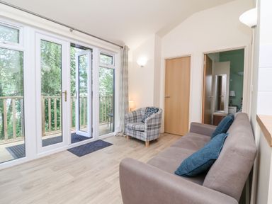A living room with a sofa with blue cushions an armchair and glass doors opening to a balcony at Chalet Log Cabin C8 in Combe Martin