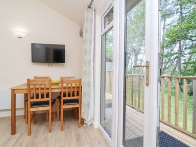 A dining area with a wooden table and four chairs next to a glass door leading to a wooden deck surrounded by trees at Chalet Log Cabin C8 in Combe Martin