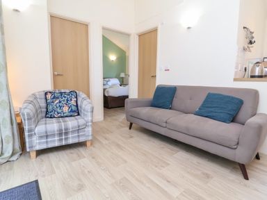 A living room with a plaid armchair and a sofa with blue cushions and open doors showing a bedroom at Chalet Log Cabin C8 in Combe Martin