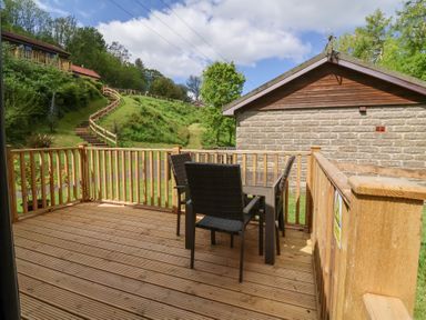 A wooden deck with a glass table and four chairs overlooking a grassy hill with stairs at Chalet Log Cabin C8 in Combe Martin