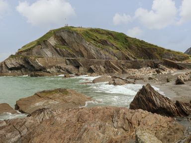 A rocky coastline with waves crashing against the shore and a grassy hill in the background at Chalet Log Cabin C9 in Combe Martin