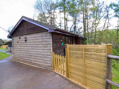 A stone and wood cabin with a wooden fence and gate surrounded by trees at Chalet Log Cabin C9 in Combe Martin
