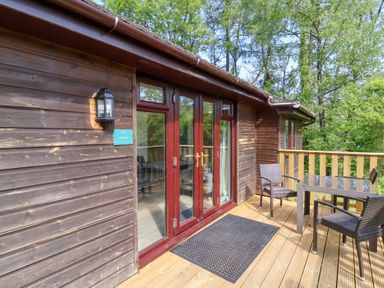 A wooden deck with glass table and three chairs outside a wooden chalet with glass doors at Chalet Log Cabin C9 in Combe Martin