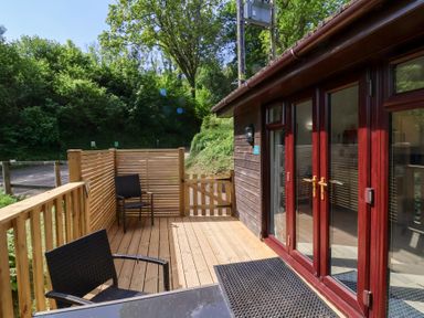 A wooden deck with two black chairs and a table next to a dark wood cabin with glass doors at Chalet Log Cabin C9 in Combe Martin
