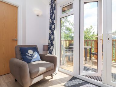 A room with a gray armchair and blue cushion next to a glass door leading to a wooden balcony with chairs at Chalet Log Cabin C9 in Combe Martin