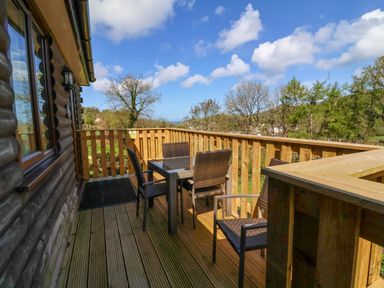 A wooden balcony with a table and four chairs overlooking trees and hills at Chalet Log Cabin L3 in Combe Martin