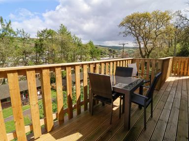 A wooden balcony with a glass table and four wicker chairs overlooking trees and houses at Chalet Log Cabin L3 in Combe Martin