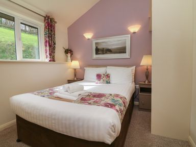 A bedroom with a double bed bedside tables lamps a window with floral curtains and a framed picture on a purple accent wall at Chalet Log Cabin L10 in Combe Martin