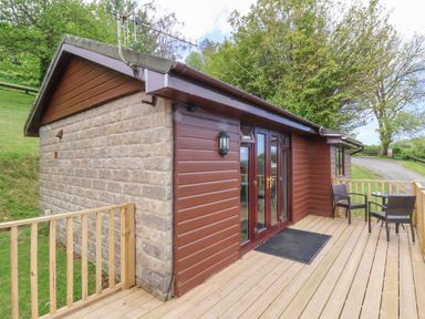 An exterior view of a small building with a wooden deck and two chairs with a table at Chalet Log Cabin L10 in Combe Martin