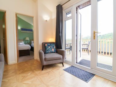 A room with an armchair next to glass doors leading to a balcony with chairs and a view of hills at Chalet Log Cabin L10 in Combe Martin