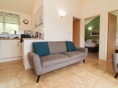 A living room with a grey sofa and turquoise pillows next to a kitchen area with appliances and a window at Chalet Log Cabin L10 in Combe Martin