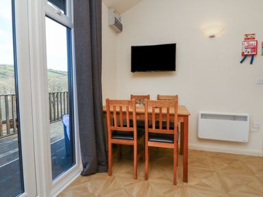 A dining area with a wooden table and four chairs next to a window and a wall-mounted television at Chalet Log Cabin L12 in Combe Martin