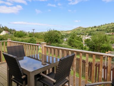 An outdoor wooden deck with a glass table and four wicker chairs overlooking green hills and houses at Chalet Log Cabin L14 in Combe Martin