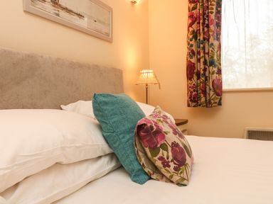 A bed with white sheets and floral and blue cushions next to a window with floral curtains at Larchwood Lodge in Combe Martin
