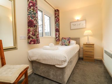 A bedroom with a single bed, floral curtains, a wooden chair with cushion, a bedside table with lamp and framed artwork at Larchwood Lodge in Combe Martin