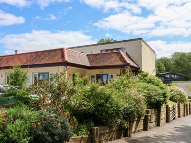 A building with a red tiled roof surrounded by bushes and flowers with a paved road and wooden fence at Larchwood Lodge in Combe Martin