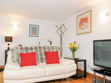 A living room with a white sofa with red cushions and a gray throw a side table with flowers a television and framed pictures on the walls at Great Brightor Cob Barn in St Kew Highway