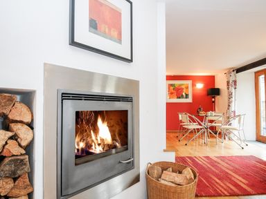 A room with a fireplace and firewood next to a dining area with a glass table and chairs at Great Brightor Cob Barn in St Kew Highway
