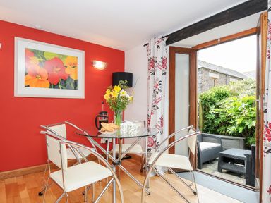 A dining area with a glass table and four chairs by an open door leading to a small patio with two black wicker chairs and a table at Great Brightor Cob Barn in St Kew Highway