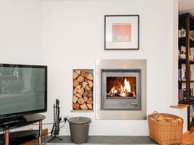 A fireplace with burning firewood and a built-in wood storage next to a television and a basket of firewood at Great Brightor Cob Barn in St Kew Highway