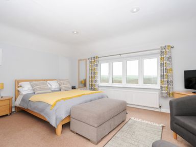 A bedroom with a bed a gray ottoman a chair and a window with patterned curtains at Great Brightor Cob Barn in St Kew Highway