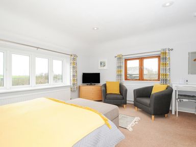 A bedroom with a bed yellow throw grey pillows two grey armchairs with yellow cushions a TV on a wooden dresser and patterned curtains at Great Brightor Cob Barn in St Kew Highway