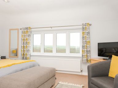 A bedroom with a window with patterned curtains a gray armchair a yellow pillow a bed with a yellow blanket and a gray ottoman at Great Brightor Cob Barn in St Kew Highway