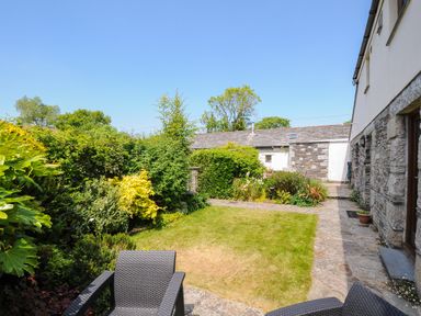 A garden with lawn surrounded by plants and shrubs and stone paved patio with chairs at Great Brightor Cob Barn in St Kew Highway