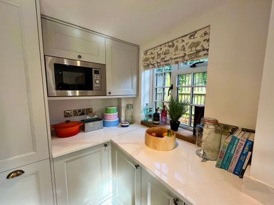 A kitchen corner with built-in microwave oven cupboards counter with fruit bowl jars and cookbooks by a window at Old Post Office in Sherborne