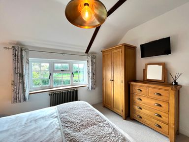 A bedroom with a bed, wooden wardrobe, wooden dresser with a mirror, a wall-mounted TV, and a window with curtains at Old Post Office Sherborne
