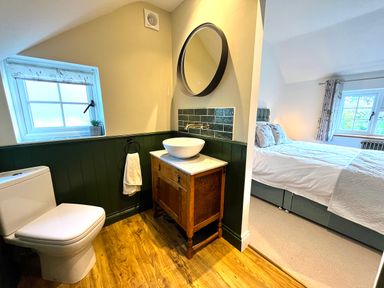 A bathroom with a toilet, wooden vanity with a round sink, round mirror, towel holder, window with patterned blind and adjacent bedroom with bed and windows at Old Post Office in Sherborne