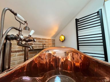 A copper bathtub with a wall-mounted light a black towel radiator and a brick wall at Old Post Office Sherborne