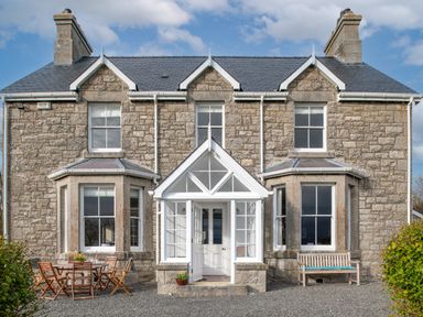 A stone house with a white porch front door a wooden table with chairs and a wooden bench at Flagstaff in Penmon near Beaumaris