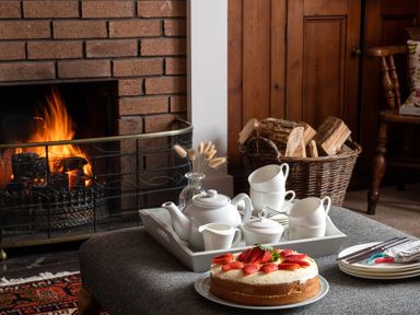 A table with a cake and a tea set near a fireplace with wood in a basket at Flagstaff in Penmon near Beaumaris