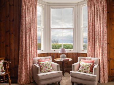 Two armchairs with patterned cushions and a round wooden table with a lamp in front of large bay windows with curtains in a wood-paneled room at Flagstaff in Penmon near Beaumaris