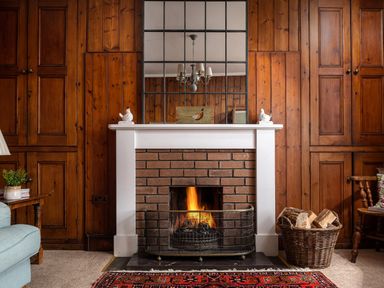 A burning fireplace with a white mantel and brick surround in a wood-paneled room with a basket of firewood and a mirror above the mantel at Flagstaff in Penmon near Beaumaris