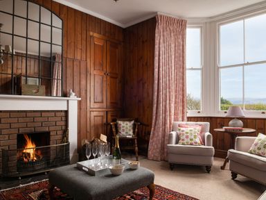 A living room with a lit fireplace wooden panel walls two armchairs with patterned cushions a wooden chair a window with curtains and a table with a lamp and books at Flagstaff in Penmon near Beaumaris