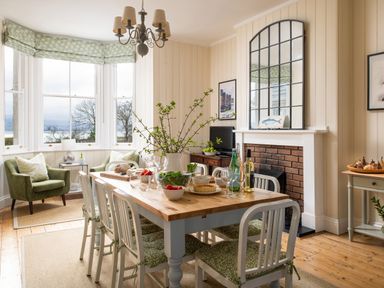 A dining room with a wooden table set with food and drinks six chairs green armchairs near bay windows and a fireplace with a large mirror above at Flagstaff in Penmon near Beaumaris