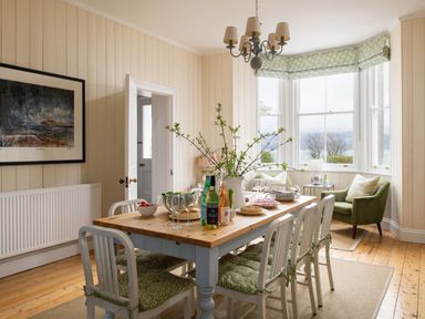 A dining room with a wooden table set with bottles, glasses, plates, and a vase of branches surrounded by white chairs with green cushions at Flagstaff in Penmon near Beaumaris