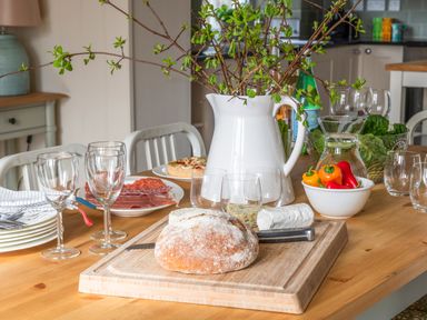 A dining table with a cutting board holding bread and cheese glasses a white pitcher with branches and fresh vegetables at Flagstaff in Penmon near Beaumaris