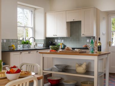 A kitchen with a wooden island table holding bread salad bottles and glasses and white cabinets with a tiled backsplash at Flagstaff in Penmon near Beaumaris