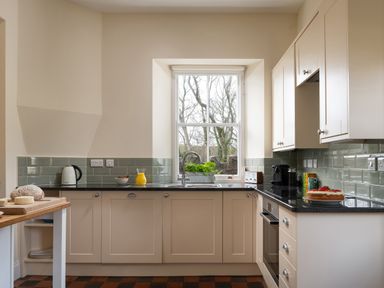 A kitchen with white cabinets black countertops a window above the sink a kettle orange juice cake and bread on surfaces at Flagstaff in Penmon near Beaumaris