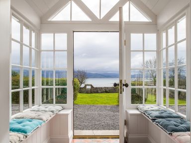 A sunroom with two cushioned benches and glass windows opening to a garden and sea view at Flagstaff in Penmon near Beaumaris