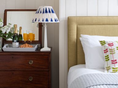 A wooden dresser with a lamp bottles of champagne glasses with orange juice and croissants next to a bed with white pillows and a decorative cushion at Flagstaff in Penmon near Beaumaris