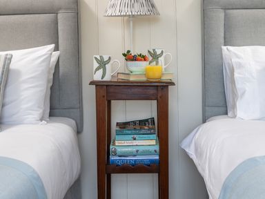 A bedside table between two beds with books mugs a bowl of fruit and a glass of orange juice at Flagstaff Penmon near Beaumaris