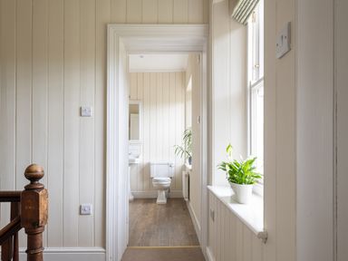 A hallway with wooden panel walls leading to a bathroom with a toilet and sink and a window with a plant on the sill at Flagstaff Penmon near Beaumaris
